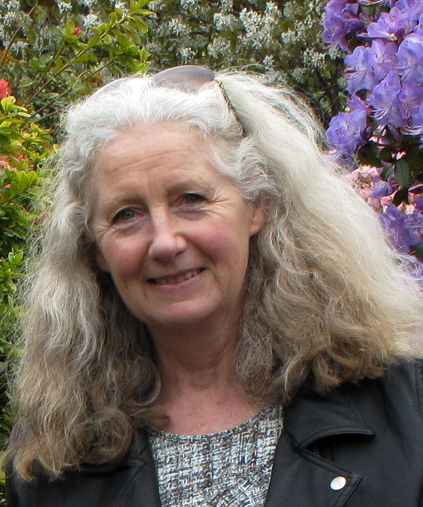 Portrait of Sharon J Yaxley, a woman with long grey hair standing in a garden.